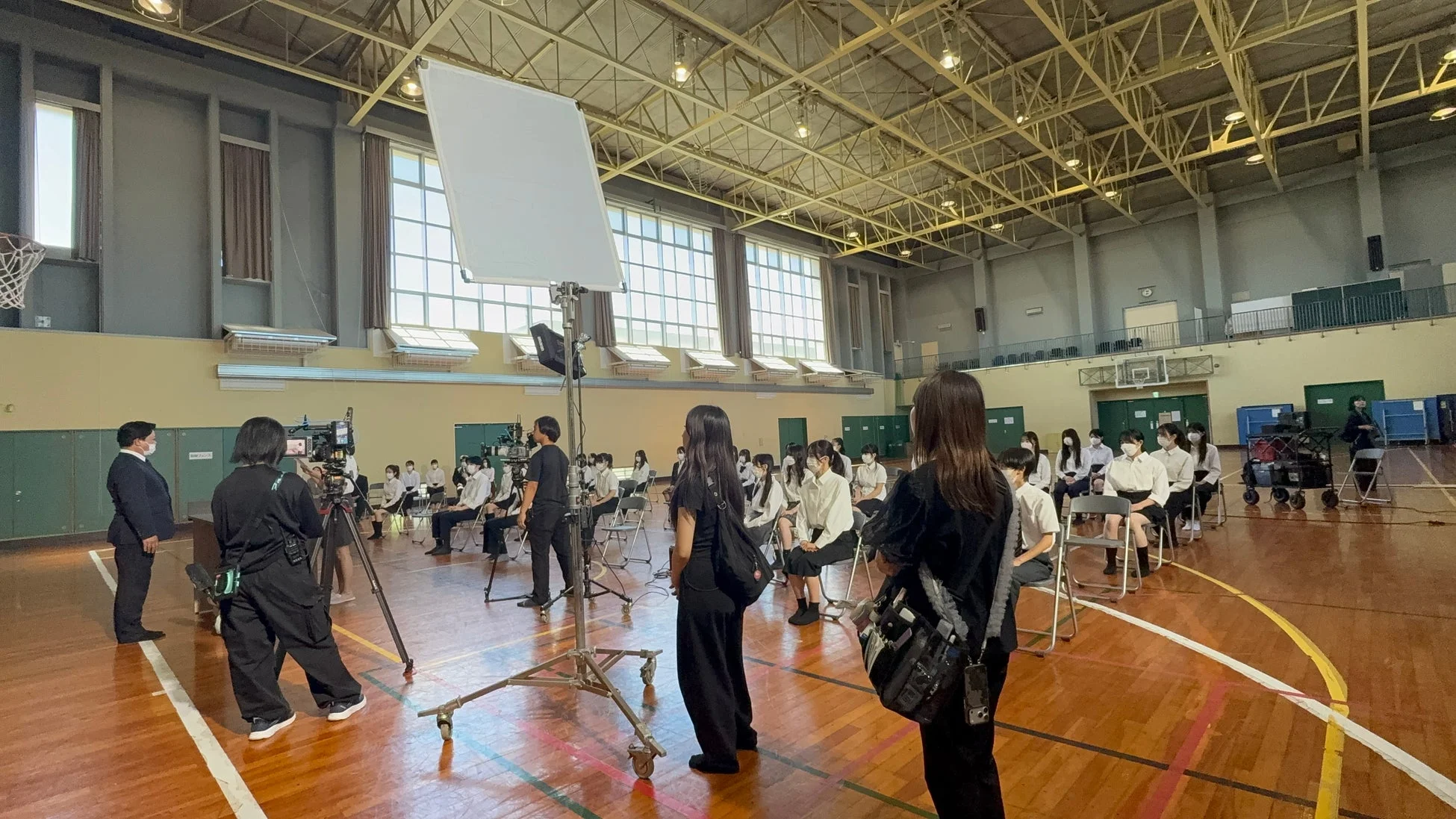 学校の体育館で映像撮影が行われている様子。複数のカメラや照明機材が設置され、制服を着た学生たちが列に座っています。