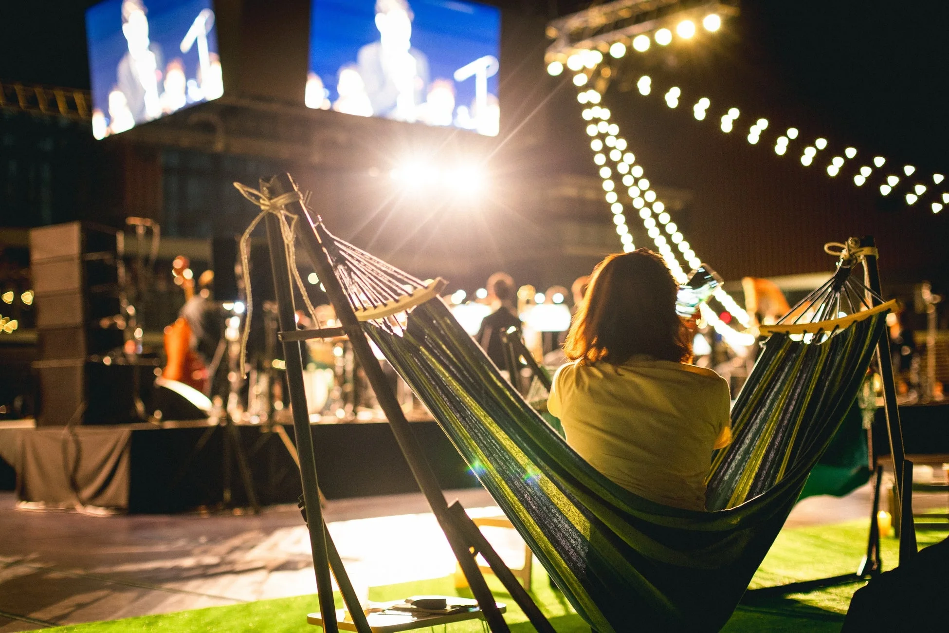 夜の屋外イベントで、ハンモックに揺られながらステージとスクリーンを眺める人の後ろ姿