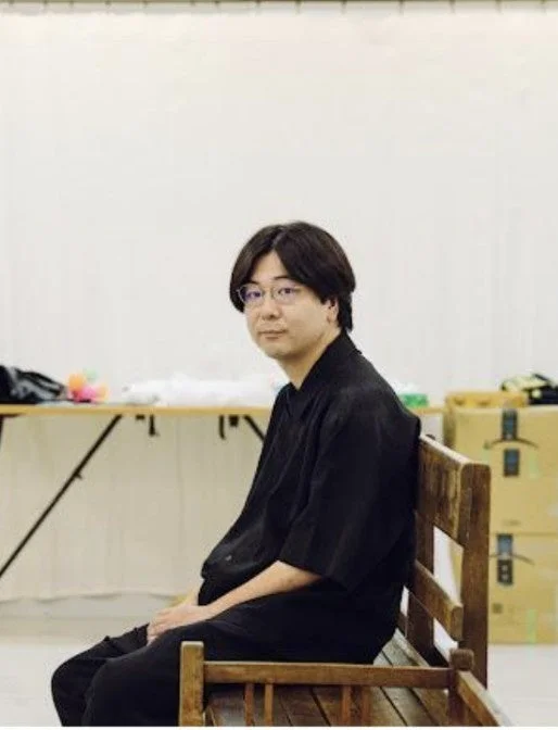 黒いシャツと眼鏡をかけたアジア系の男性が木製のベンチに座り、カメラの方を向いている画像