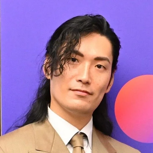 紫色の背景と赤い円を背に、長くてカールした黒髪の男性がカメラを見つめているポートレート