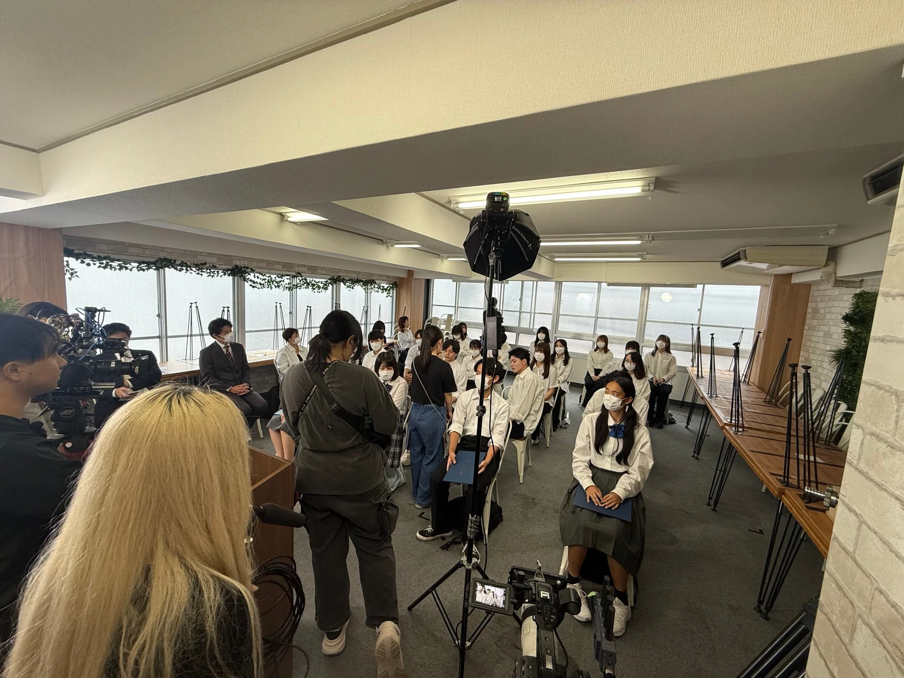 カメラや照明が設置された室内で、制服姿の学生たちが着席し、撮影やインタビューを受けている様子。