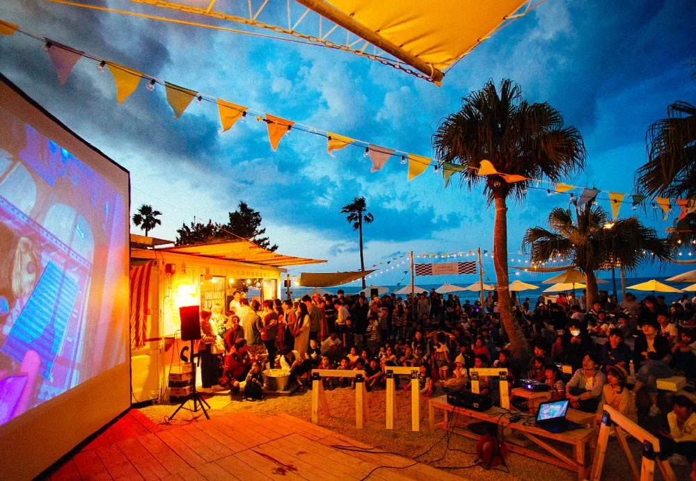 夕暮れ時、ビーチのような場所で多くの人々が屋外スクリーンに映し出された映画を鑑賞