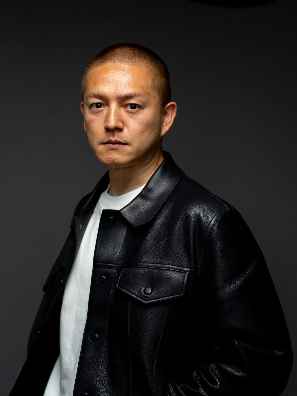 暗い背景を背に、坊主頭の男性が黒いレザージャケットと白いTシャツを着用し、カメラをまっすぐ見つめているポートレート写真