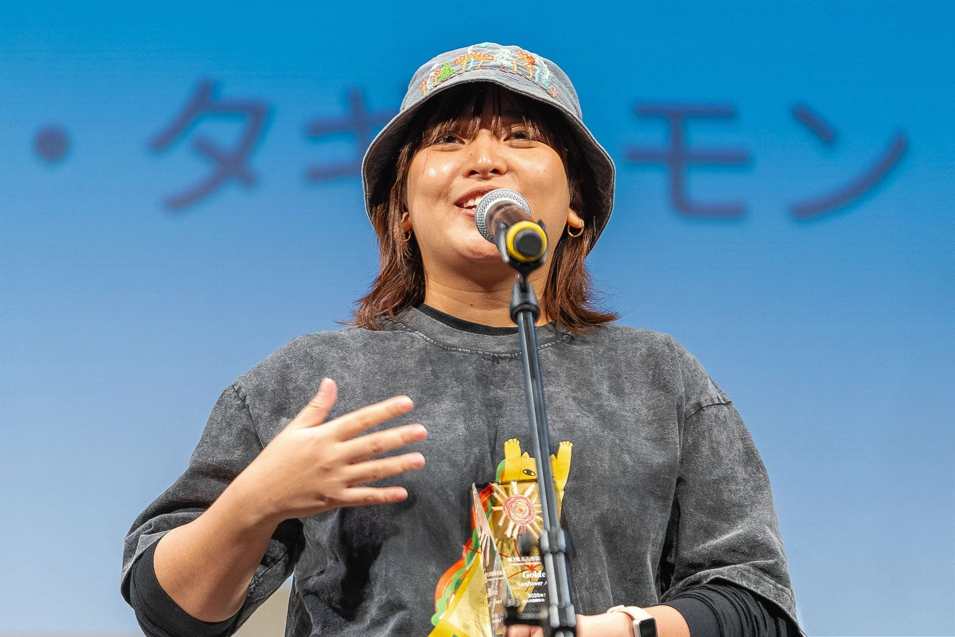 バケットハットをかぶった女性がマイクに向かってスピーチしており、右手にはトロフィーらしきものを持っています